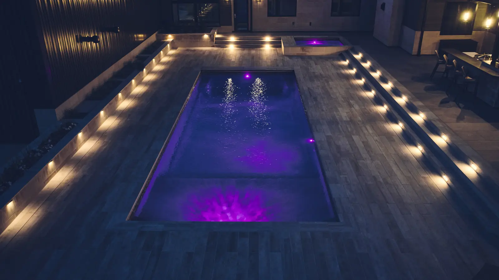 Looking down a purple-lit pool at night with step lighting along both sides of the deck