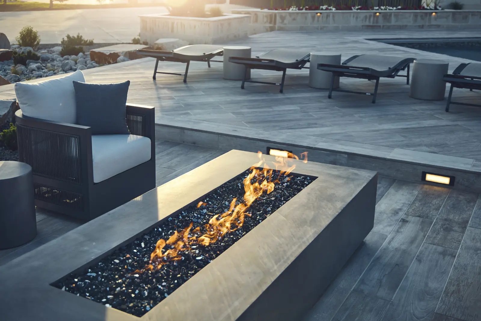 Flames in a linear concrete fire pit beside outdoor armchairs on the paver deck
