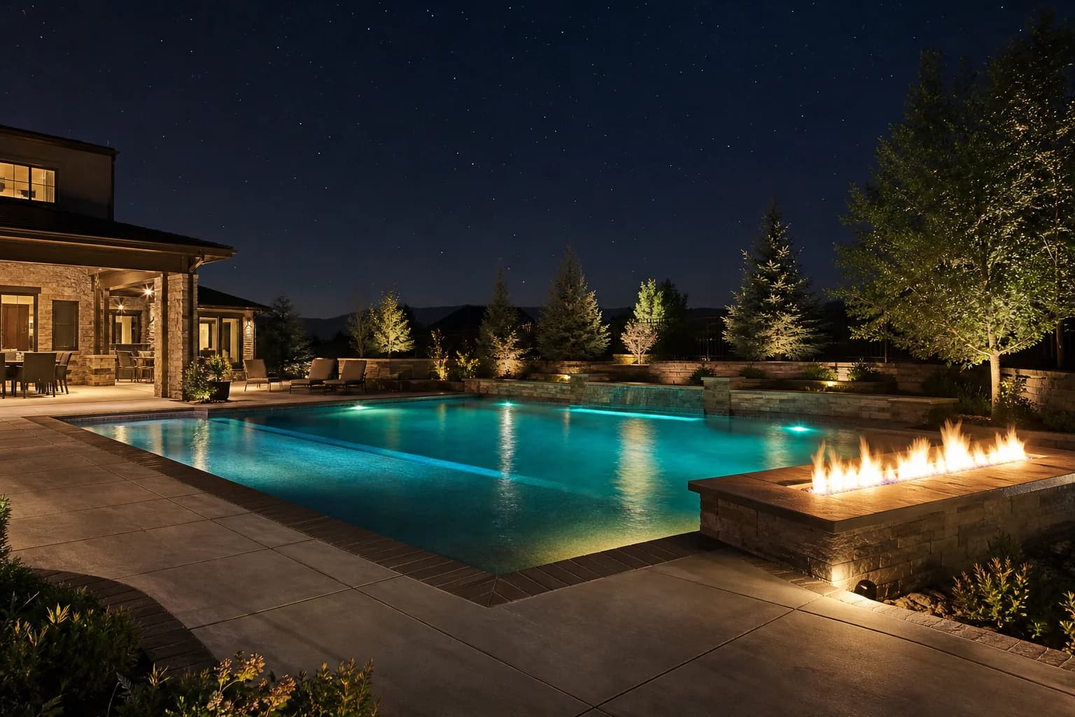 Night-lit swimming pool with linear gas fire feature on stone pedestal and paver deck surround
