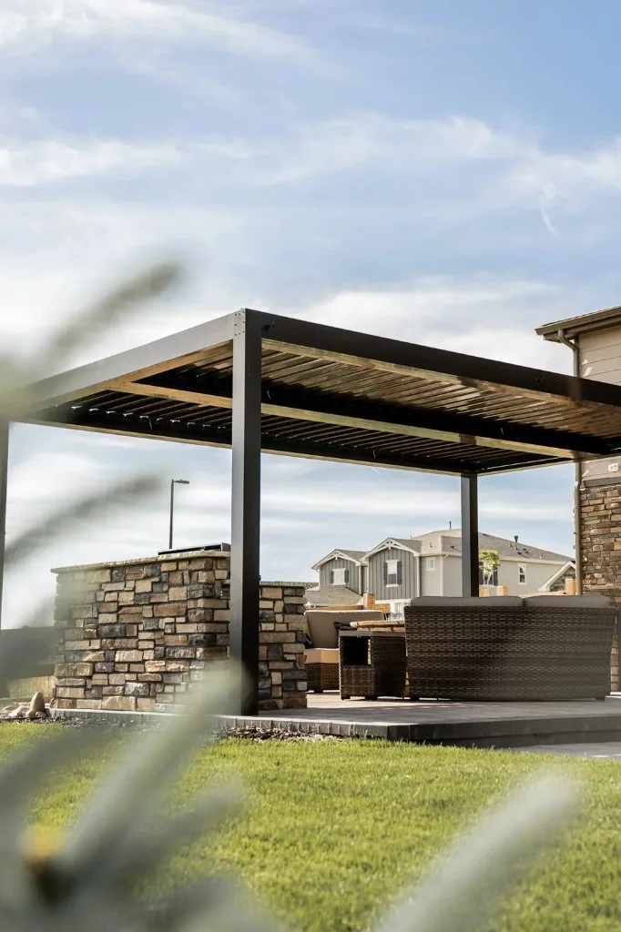 Black louvered pergola over stone-veneered outdoor kitchen island, wicker seating, and large-format patio pavers, integrated shade, cooking, and hardscape in one composition.