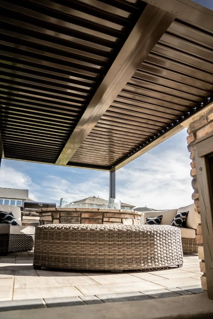 Louvered bronze patio cover over light tan pavers, circular stone fire pit with glass wind guard, and cushioned wicker seating, adjustable shade over a true outdoor room.