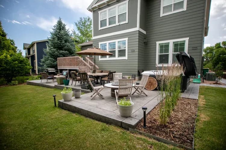 Wide backyard view: two-tier stamped concrete patio, dining under umbrella, lounge seating, hot tub, grill under deck, and lawn, Westminster Sorensen project.